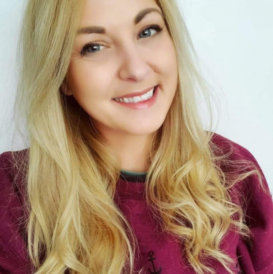 The image shows a smiling woman with long blonde hair cascading over her shoulders. She has light skin and wears a maroon sweater with an anchor symbol visible on the chest area. Her bright and friendly expression is highlighted by her clear, blue eyes and a natural makeup look. The background is plain white, emphasizing her features and warm demeanor.