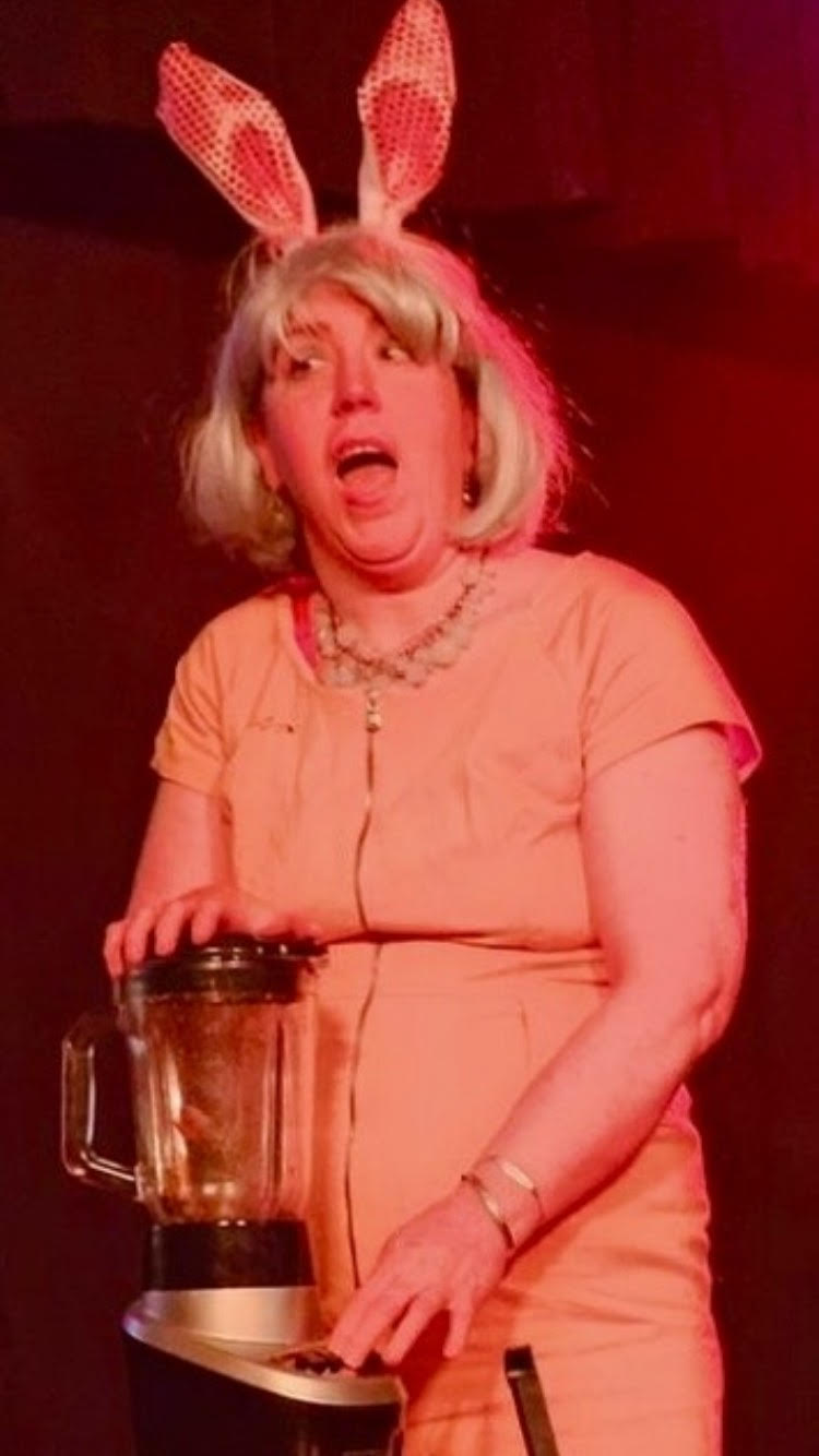 The image shows a performer mid-act, dressed in a peach outfit with a bold necklace and bunny ears, holding onto a blender. The lighting creates a dramatic and comedic atmosphere, likely capturing a humorous moment from a live show. The person’s expression adds to the lively and engaging nature of the performance, suggesting a playful or theatrical context.