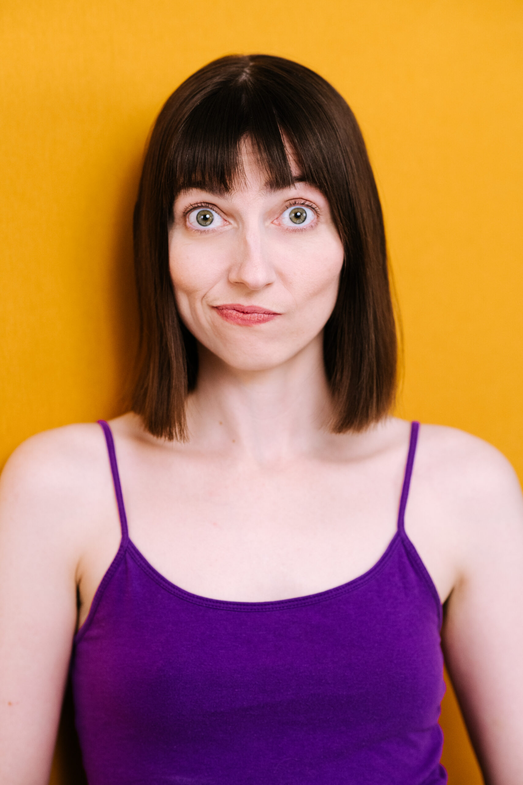 This is a portrait of a young woman with straight brown hair cut in a neat, shoulder-length bob and bangs. She has strikingly large, light green eyes and a small, subtle smile. Her expression is friendly and open, with a slight air of curiosity or amusement. She is wearing a vibrant purple sleeveless top, and the background is a solid, cheerful yellow, which creates a striking contrast with her outfit and hair, making her features stand out prominently. The overall tone of the photo is bright and inviting.