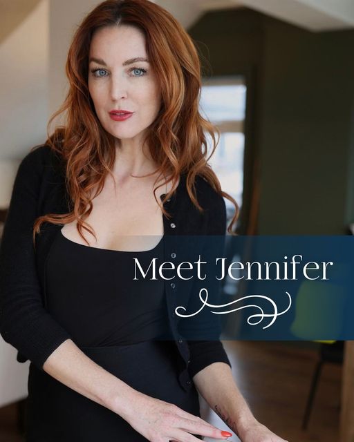 This image features a professional portrait of a woman identified as "Jennifer." Here's a detailed description: Description: Subject: The woman has long, wavy, auburn hair cascading past her shoulders. She is wearing a sleek black dress paired with a black cardigan, giving her a sophisticated and elegant look. Her makeup is natural yet polished, with bold red lipstick adding a touch of glamour. Pose and Expression: Jennifer is standing with a confident and poised demeanor, looking directly at the camera. Her expression is warm and inviting, exuding approachability and professionalism. Background: The setting appears to be an indoor space, possibly a modern home or studio. The background is softly blurred, showing a hint of large windows and minimalistic decor. Text: The words "Meet Jennifer" are overlaid on the lower right portion of the image in elegant typography, accompanied by a decorative flourish. This photo conveys a professional and approachable image, likely intended for a personal introduction or promotional purpose.