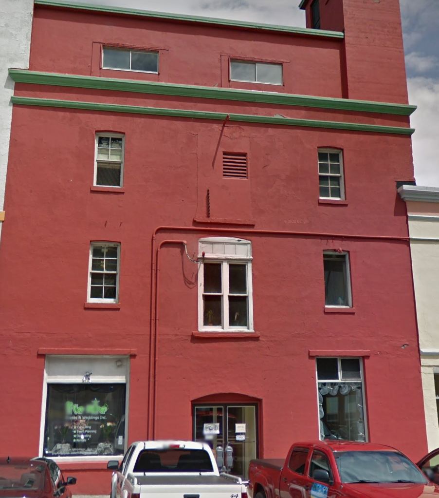 This is an image of a tall, red brick building with green trim along the edges of the roof and structural details. The building appears to be in an urban setting with vehicles parked in front, including a white truck and a red pickup. The ground floor features commercial windows, with one displaying signage for a business related to events or weddings. The upper floors have several windows, some of which are small and rectangular. The exterior shows signs of wear, with patches of uneven paint and exposed brickwork. The building has a distinct architectural charm, possibly a historical or repurposed structure.