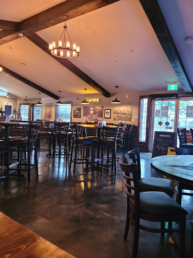 This is an interior photograph of a cozy and modern pub or restaurant. Here are the key details: Description: Seating and Layout: The room features several round and square wooden tables surrounded by cushioned chairs. High-top tables with bar stools are scattered across the space, suggesting a casual and social atmosphere. Lighting: Warm and inviting light fills the room, coming from chandeliers with a rustic, wagon-wheel design, and pendant lamps. Natural light filters in through the large windows and a glass door on one side. Decor: Exposed wooden beams run across the ceiling, adding to the rustic charm. A chalkboard menu is visible near the entrance, adding a personal touch. Wall art and signs, including a prominent "Corona" sign, decorate the walls, enhancing the relaxed vibe. Flooring: The flooring is polished concrete, lending a sleek and contemporary feel. This space seems well-suited for gatherings, casual dining, or enjoying drinks in a comfortable and welcoming environment.