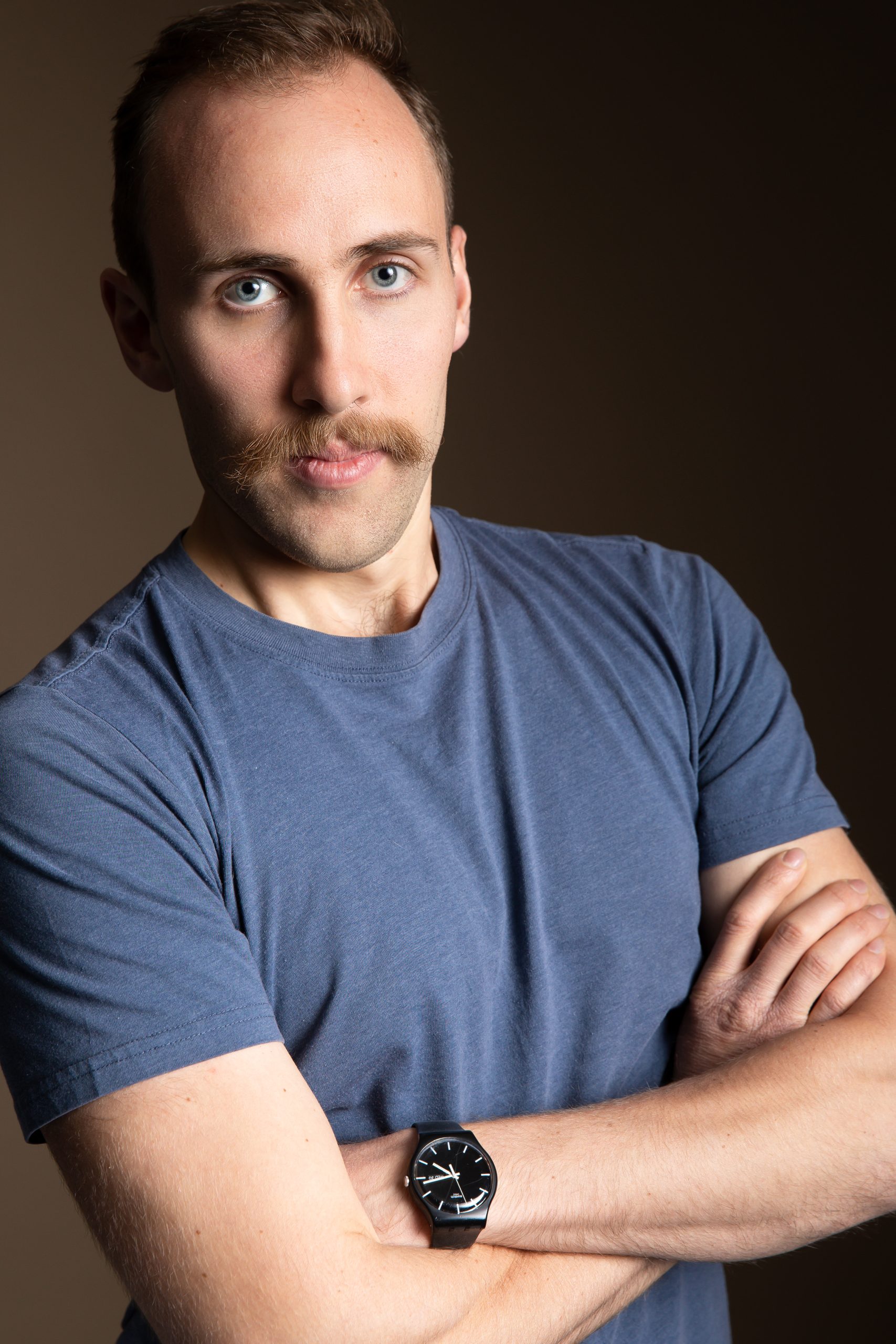 This is a professional portrait of a man with light skin, blue eyes, and a distinctive mustache. He has a confident expression, with his arms crossed, and is wearing a fitted blue T-shirt and a black wristwatch with a sleek design. The background is a simple, neutral-toned setting, giving the photo a polished, studio-like appearance. The lighting is soft and even, highlighting his facial features and giving the image a clean, professional feel.