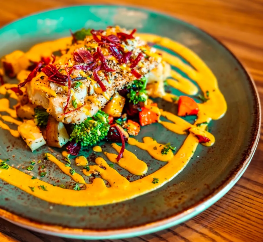 Description of the Image: This is a beautifully plated gourmet dish featuring a perfectly seasoned and crusted white fish fillet on a bed of roasted vegetables. The fish has a golden, herb-infused crust and is topped with crispy, thinly sliced red onions or beets. The plate is decorated with a vibrant yellow sauce, drizzled artistically in swirls and streaks. The vegetables beneath the fish include broccoli, carrots, and cubed potatoes, all appearing to be roasted to perfection, adding a mix of green, orange, and golden hues. The dish is served on a rustic turquoise ceramic plate, which contrasts beautifully with the warm colors of the food. The background is a wooden table, adding to the cozy, high-end dining feel. Fresh herbs like parsley or dill are sprinkled over the plate, enhancing the dish’s visual appeal and freshness. This dish looks like something you’d find at a fine dining restaurant or a high-end seafood bistro!