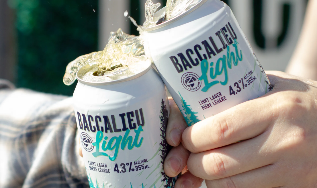 Description of the Image: This vibrant action shot captures two hands holding and clinking open cans of "Baccalieu Light" beer, creating a splash of liquid mid-air. The cans are white with teal and black lettering, featuring a forest silhouette design, giving a fresh and crisp aesthetic. The beer is labeled as "Baccalieu Light", a light lager with 4.3% alcohol content, packaged in 355ml cans. The branding includes a circular logo with the name "Baccalieu Brewing", indicating the brewery's identity. The splashing effect from the opened cans adds a sense of excitement and celebration. The background is slightly blurred, showing an outdoor setting with a mix of green and neutral tones, reinforcing the refreshing and social nature of the beer. This image is dynamic, refreshing, and energetic, likely promoting Baccalieu Light as a go-to drink for social gatherings and outdoor enjoyment.