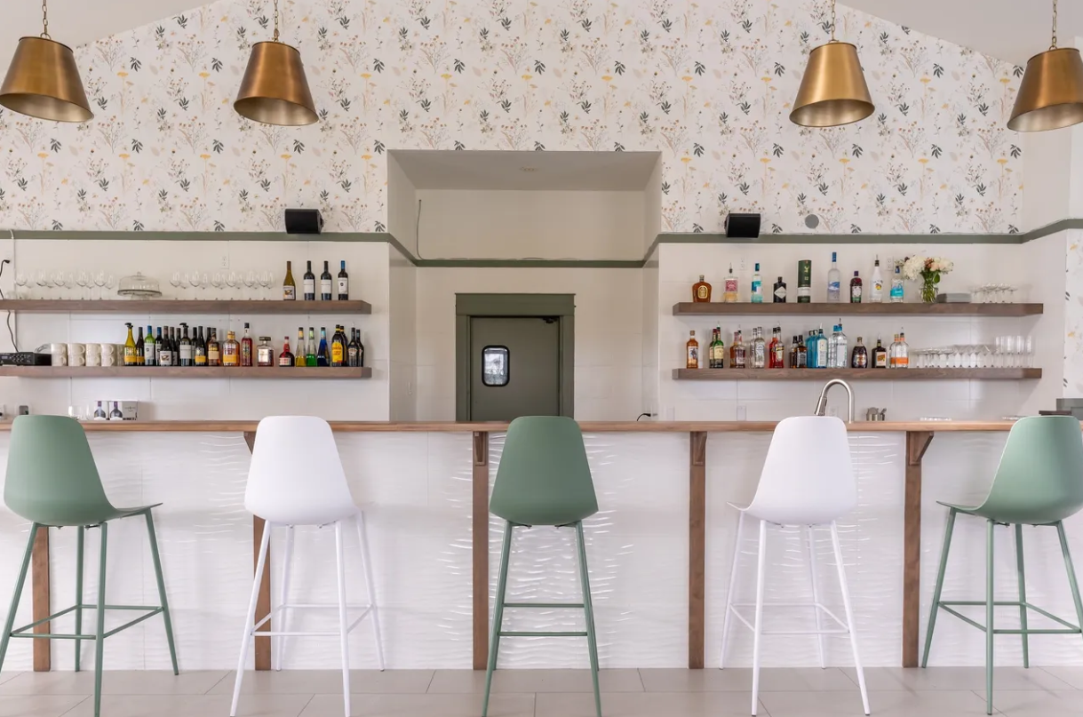 This image showcases a beautifully designed bar interior with a clean and modern aesthetic. The bar features a white counter with a wood-accented top, lined with four high barstools in alternating shades of white and muted green. The back wall is decorated with elegant floral wallpaper, and wooden floating shelves hold an array of liquor bottles, glassware, and decorative items. Above the bar, three large brass pendant lights add warmth and sophistication to the space. The center of the bar features a doorway with a small window, leading to what appears to be the kitchen or storage area. The overall ambiance is bright, airy, and inviting, with a minimalist yet stylish approach to design.