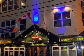 This image shows the exterior of Bridie Molloy's, a well-known pub and lounge in St. John's, Newfoundland. The entrance features a black awning with the pub’s logo in the center, flanked by two flags—a Canadian flag and an Irish flag—which reflect the venue’s Irish pub identity. The building's facade is illuminated by blue and yellow lights, giving it a warm and inviting nighttime atmosphere. Bridie Molloy's is a popular spot for live music, stand-up comedy, and social gatherings, making it a great venue for events like the "Two Idiots Stand-Up Comedy" show on March 7th. The pub's traditional Irish aesthetic and cozy ambiance make it a favorite among both locals and visitors. 🍀🍻