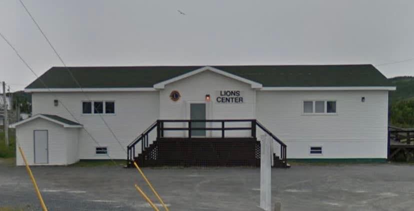 The image shows the Lions Center, a single-story, rectangular community hall with a white exterior and dark green roof. The front facade features a symmetrical layout, with a central entrance accessed by a wide, raised wooden deck and staircase painted dark brown. The entrance is framed by a triangular peaked roof, giving it a clean and welcoming appearance. Two small Lions Club crests are mounted beside the front door, along with the text “LIONS CENTER” in bold black letters. There are multiple rectangular windows on both sides of the building, allowing natural light inside. To the left of the main structure is a small white outbuilding with a sloped roof, possibly a storage shed or utility room. The building sits on a gravel lot with visible yellow parking lines and is surrounded by light vegetation in the background, suggesting a rural or small-town setting. The overall appearance is tidy, functional, and typical of a well-maintained community or service club building in Newfoundland and Labrador.