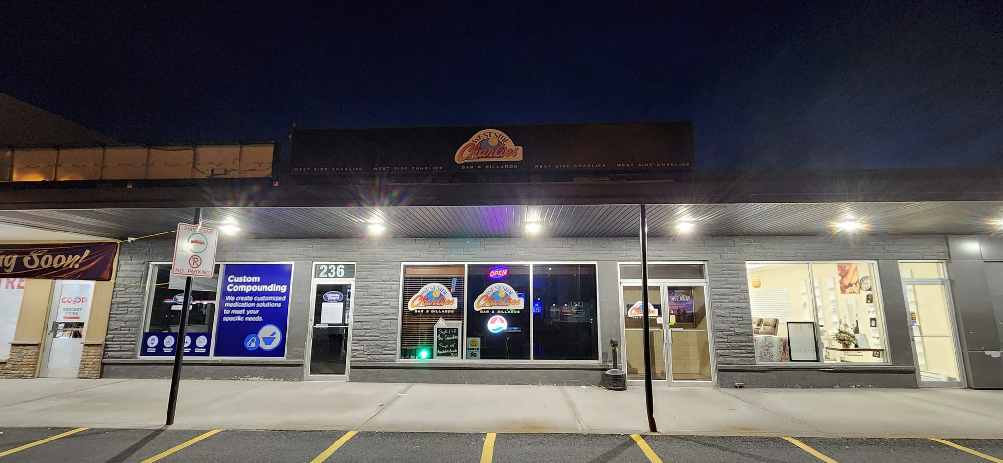 This is a nighttime view of West Side Charlie's, a bar and billiards establishment. The location appears to be: 📍 236 Memorial Drive, likely in Clarenville, NL, based on your recent uploads and events. Here are a few notable visual details: The signage clearly says "West Side Charlie's Bar & Billiards", lit with neon. There's a Co-op grocery store nearby and a pharmacy with “Custom Compounding” services, making this part of a small retail plaza. The entrance has signage for upcoming events and promotions posted in the windows.
