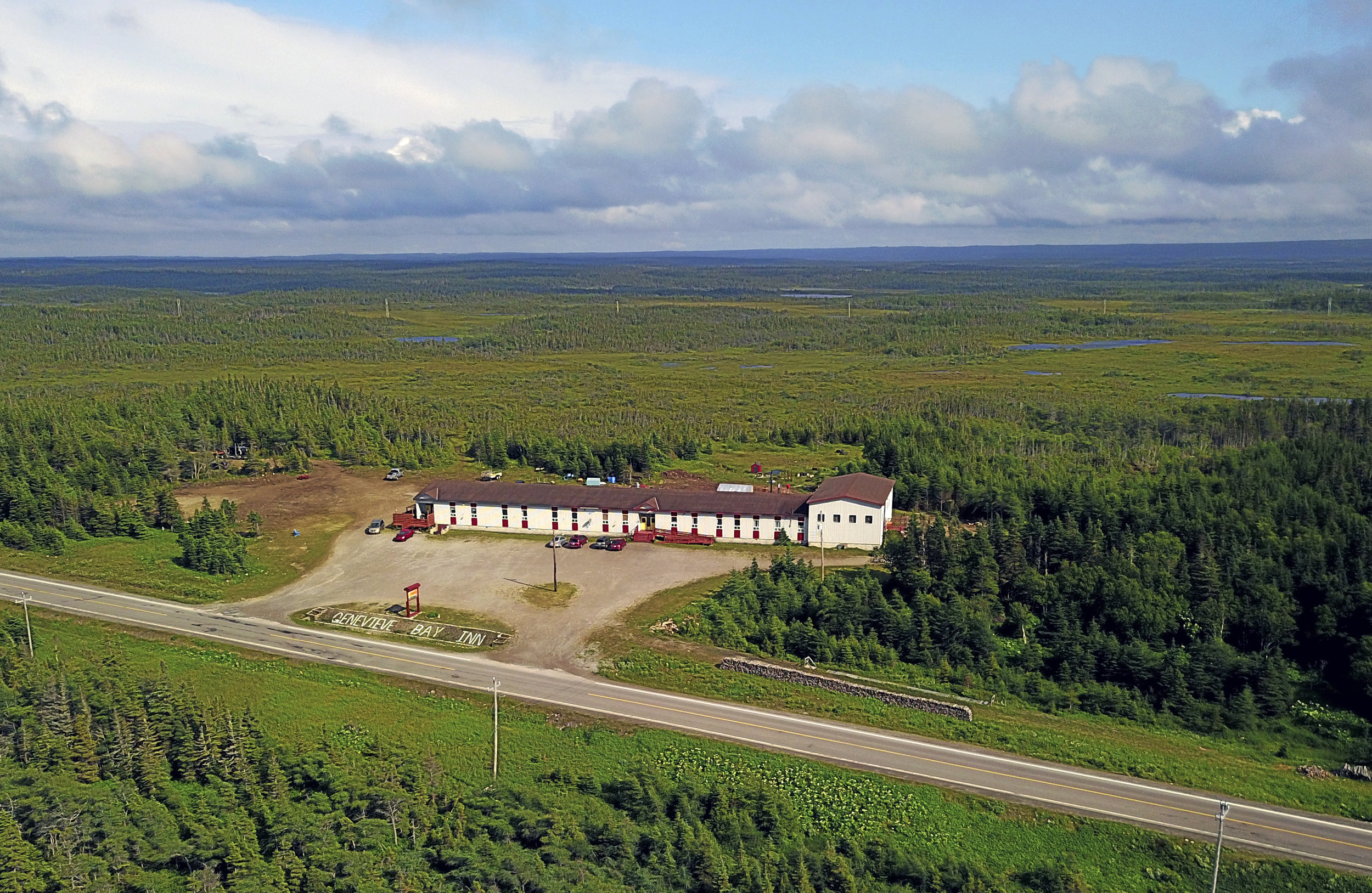 This aerial image captures the Genevieve Bay Inn, a remote and spacious hotel nestled in the natural beauty of St. Barbe, Newfoundland and Labrador. Located just off the main highway, it offers both isolation and accessibility, ideal for travelers, ferry passengers, and event-goers alike. Venue Highlights: The large, single-level main building is set back from the road with a clearly marked sign reading “GENEVIEVE BAY INN.” The front lot has ample parking space for guests and event attendees. Surrounded by thick boreal forest and expansive wilderness, the location provides a serene and scenic backdrop for any event. It’s connected to Thirsty’s Lounge, a known local bar and live entertainment spot inside the inn. This location is well-suited for rural comedy nights, live music events, or intimate gatherings where nature meets nightlife.