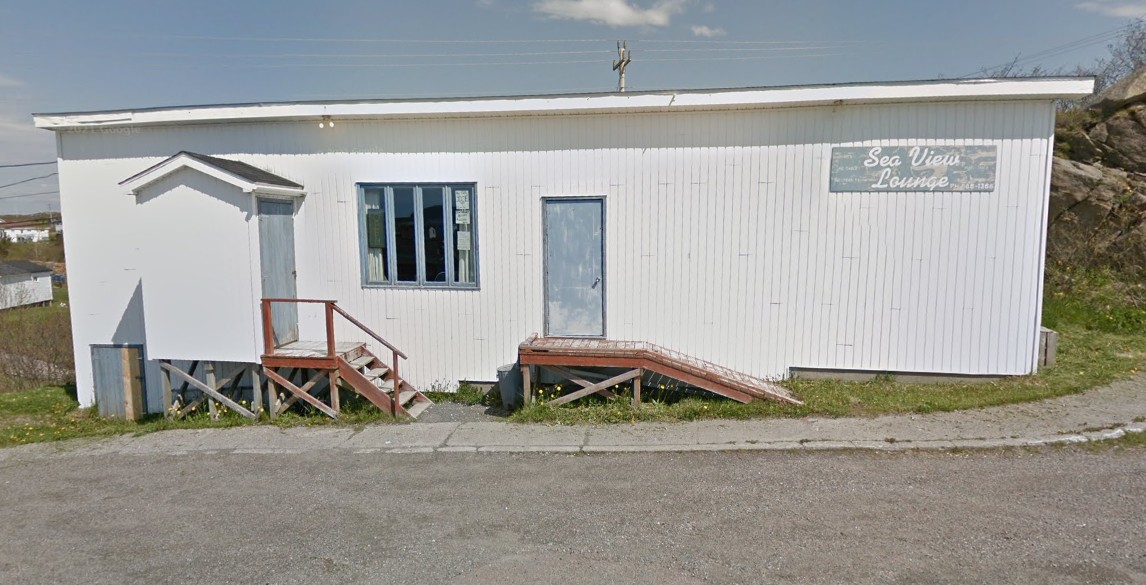 The image shows the Sea View Lounge, a small, modest venue with a white wooden exterior and a flat roof. The building has a rectangular, elongated layout and sits on a slight elevation next to a rocky slope. On the left side, there’s a small enclosed porch with a pitched roof and a basic wooden staircase leading up to a door. In the center, there’s a set of three-panel windows and a secondary entrance with a wooden ramp, suggesting a nod to accessibility, though it appears aged and possibly steep by modern standards. Mounted above the door on the far right is a weathered sign reading: Sea View Lounge Ph: 886-3166 The exterior gives off a charming, no-frills, rural Newfoundland vibe—likely a well-loved local hangout for live music, community events, and comedy nights. The lounge appears to serve a tight-knit community and may have that classic “everyone-knows-your-name” feel inside.
