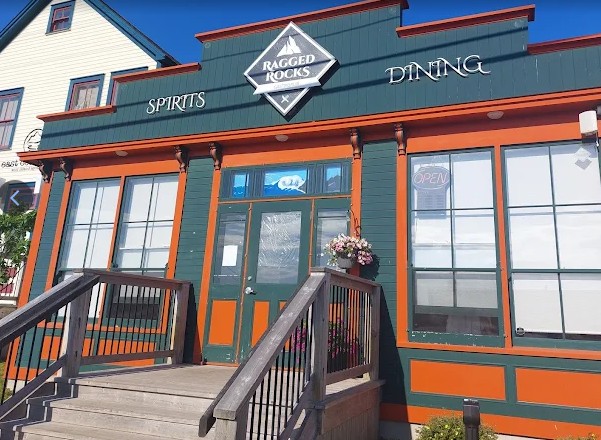 This image shows the Ragged Rocks Gastropub, a stylish and inviting venue located in Bonavista, Newfoundland and Labrador. The building has a distinct character, with deep green siding accented by burnt orange trim, giving it a warm, heritage-inspired look that fits perfectly in Bonavista's historic waterfront area. Key features: The signage reads "Ragged Rocks" with bold letters and a diamond-shaped logo above the entrance. The words "SPIRITS" and "DINING" are written in elegant script on either side of the logo. The entryway features a set of wooden stairs with sturdy railings, leading up to a glass-paneled double door. There are large front windows that likely offer a view of the street or water, adding to the cozy atmosphere inside. A small "OPEN" sign is visible in one of the windows, suggesting it's a welcoming and frequented place for locals and tourists alike. This venue is well-suited for comedy shows, live music, and dining experiences with character