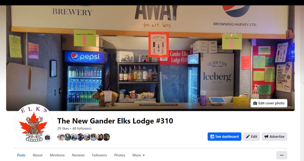This is the bar area inside the newly renovated Gander Elks Lodge #310, where our “Laughs at the Elks” showcase on April 20 will take place. A long service counter stretches across the frame, backed by chilled beverage coolers stocked with Pepsi products on the left and Iceberg wine coolers on the right. Between them, shelves display a handful of spirits and grab-and-go snacks. Above the counter, signage from local sponsors (including our brewery partners) hangs on a supportive beam, with clear “Order Here” and “Pay Here” arrows taped to guide patrons. Brightly colored posters and notices line the sidewalls—everything from upcoming event flyers to reminders about reusable cups—giving the space a cozy, community-driven vibe. In front of the counter is a small table area where you can catch up with friends before heading into the performance hall. It’s a friendly, no-frills venue that’s perfect for an intimate comedy night.