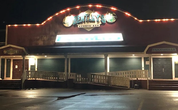 This image shows the exterior of Maxwell’s & Bentley’s, a well-known venue in Goose Bay, NL. The facade is warmly lit at night, creating a welcoming glow that highlights the building’s curved roofline and signage. The entrance features a wide, accessible ramp alongside traditional stairs, with railings guiding guests to the front doors under soft canopy lighting. The left and right entrances are branded separately—Bentley’s Sports Bar on one side, Maxwell’s on the other—hinting at multiple spaces within the building. This venue is perfect for live performances, combining the comfort of a community bar with the capacity and layout of a proper event space. Ideal for a rowdy, high-energy comedy night.